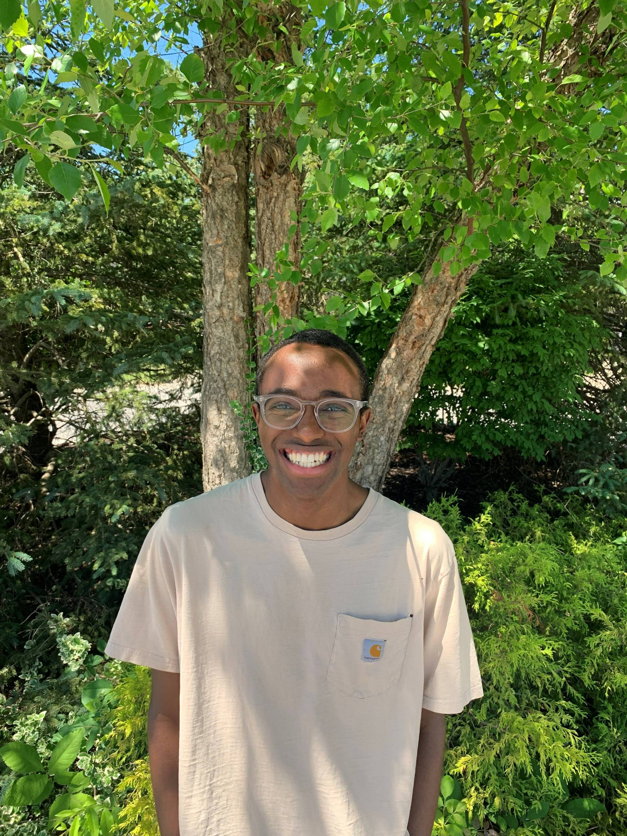 A student stands in front of a lush green background smiling.